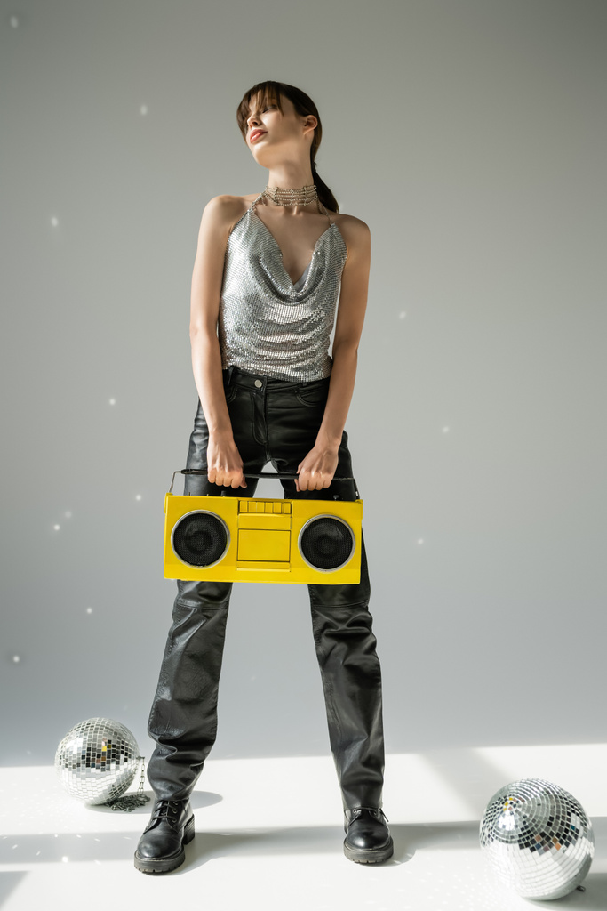 full length of woman in trendy top and leather pants holding boombox near disco balls on grey  - Photo, Image