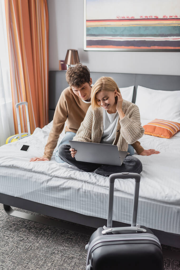 smiling woman sitting on hotel bed with laptop near young boyfriend and suitcase on foreground - Photo, Image