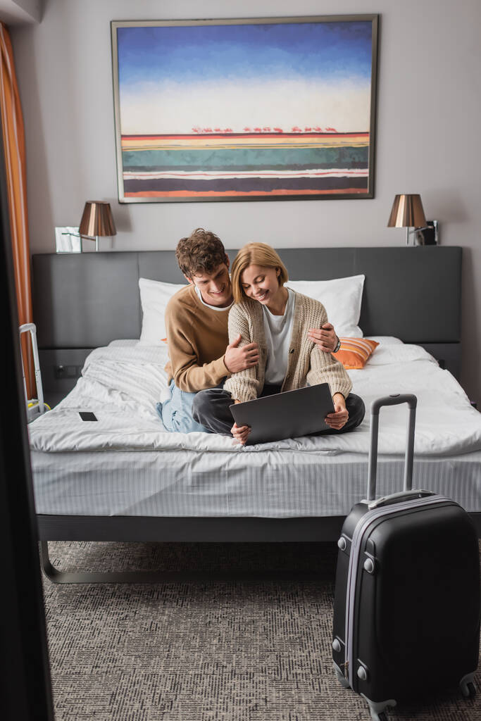 happy woman sitting with laptop near smiling boyfriend on bed in modern hotel - Valokuva, kuva
