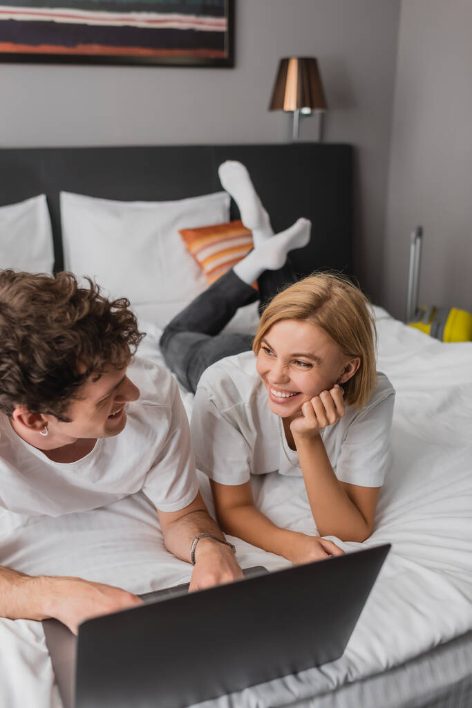 cheerful blonde woman looking at boyfriend near laptop on bed in hotel - Zdjęcie, obraz