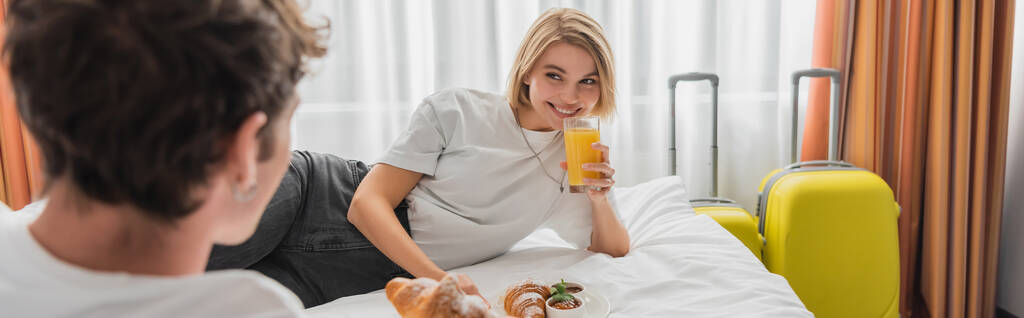ευτυχισμένη γυναίκα με ένα ποτήρι χυμό πορτοκάλι κοιτάζοντας θολή φίλο στο κρεβάτι στα διαμερίσματα του ξενοδοχείου, πανό - Φωτογραφία, εικόνα