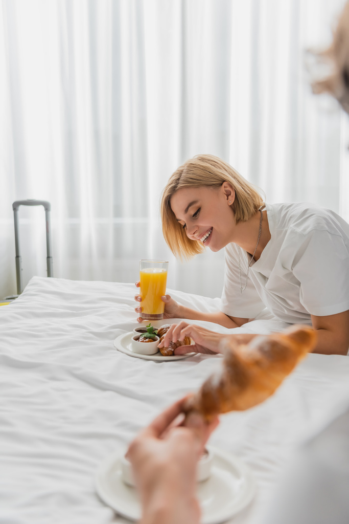 веселая блондинка завтракает апельсиновым соком и круассаном на кровати отеля рядом с размытым парнем - Фото, изображение