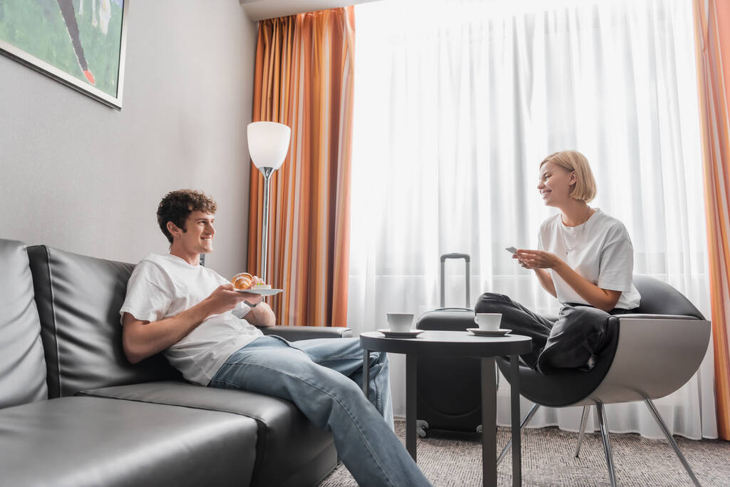 young woman with smartphone sitting in armchair near boyfriend with croissant in modern hotel suite - Foto, afbeelding