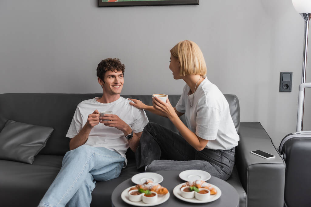 blonde woman with cup talking to smiling boyfriend near croissants and jam with chocolate paste on coffee table - Foto, imagen