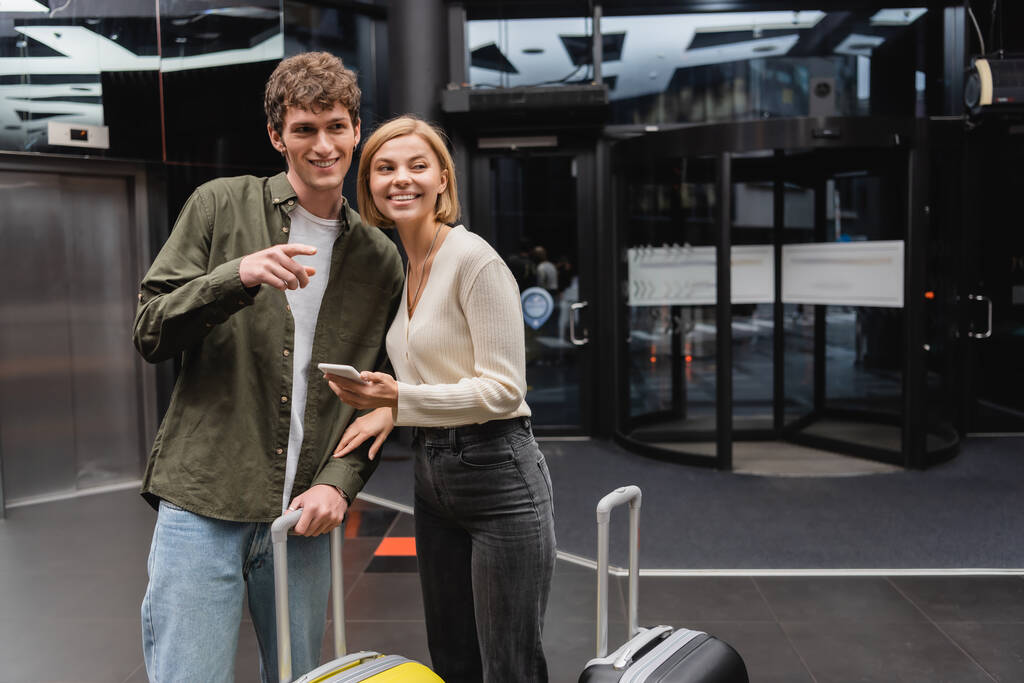 young man pointing with finger and looking away near smiling girlfriend and travel bags in hotel - Foto, Imagen