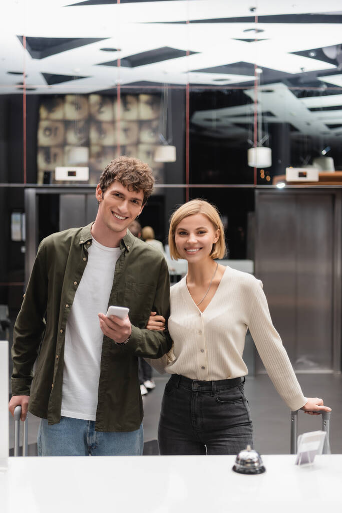 blonde woman and brunette man with smartphone smiling at camera near reception in modern hotel - Photo, image