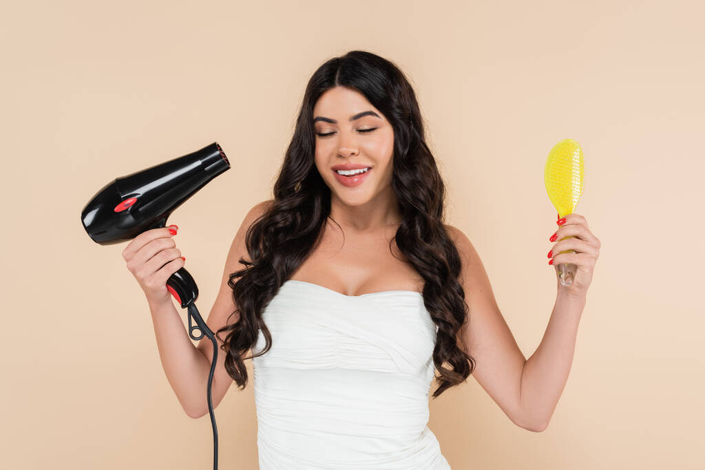 Smiling brunette woman holding hairbrush and hairdryer isolated on beige  - Photo, image