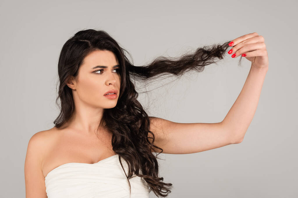 displeased woman in white top looking at damaged and tangled hair isolated on grey - Foto, Imagen