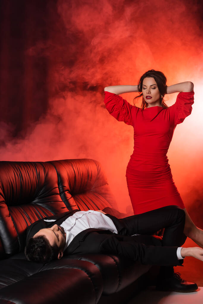 brunette woman with closed eyes seducing boyfriend in formal wear lying on couch on red with smoke   - Fotó, kép