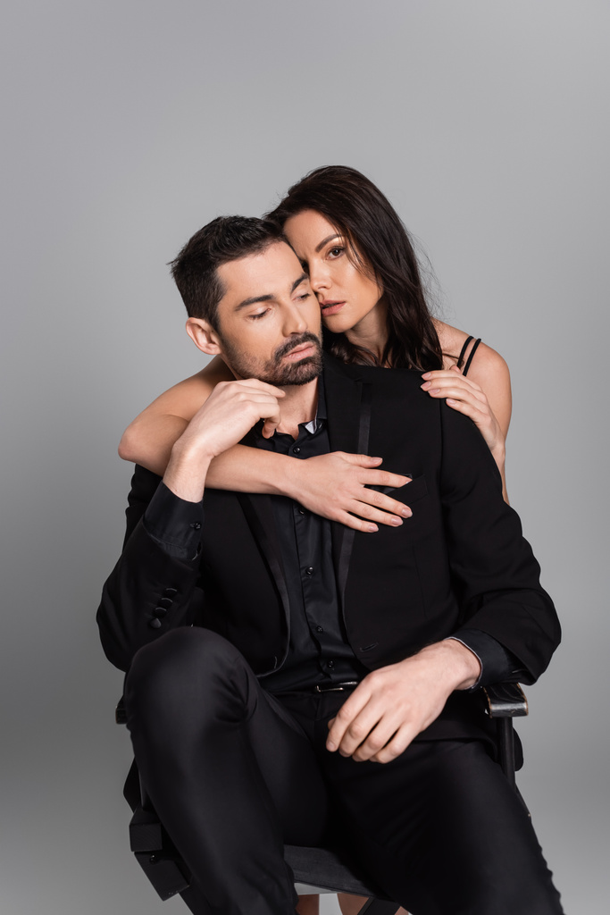 brunette woman hugging bearded man in black suit sitting isolated on grey  - Photo, image