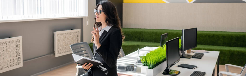 brunette businesswoman in eyeglasses holding notebook while thinking near desk with monitors in office, banner - Foto, afbeelding