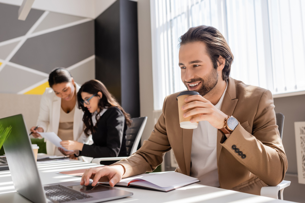 улыбающийся менеджер с напитком на вынос глядя на ноутбук рядом с мультиэтнических деловых женщин на размытом фоне - Фото, изображение