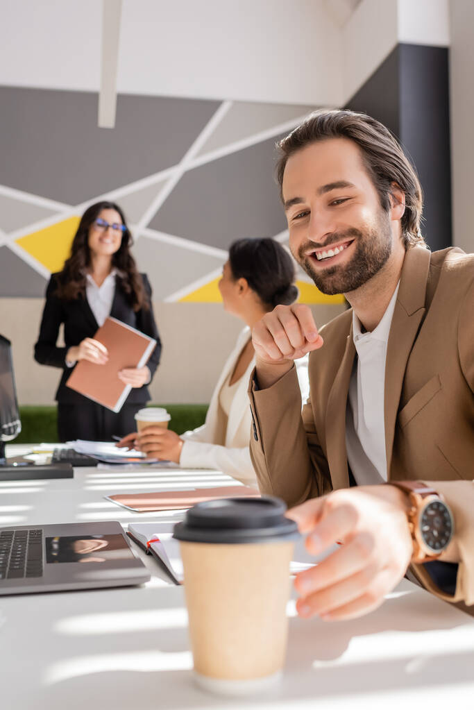 smiling manager reaching takeaway drink while interracial businesswomen talking on blurred background - Foto, Bild