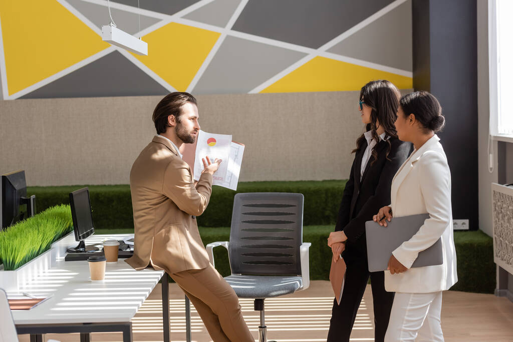 боковой вид бизнесмена, показывающего графики мультикультурным коллегам в современном офисе - Фото, изображение