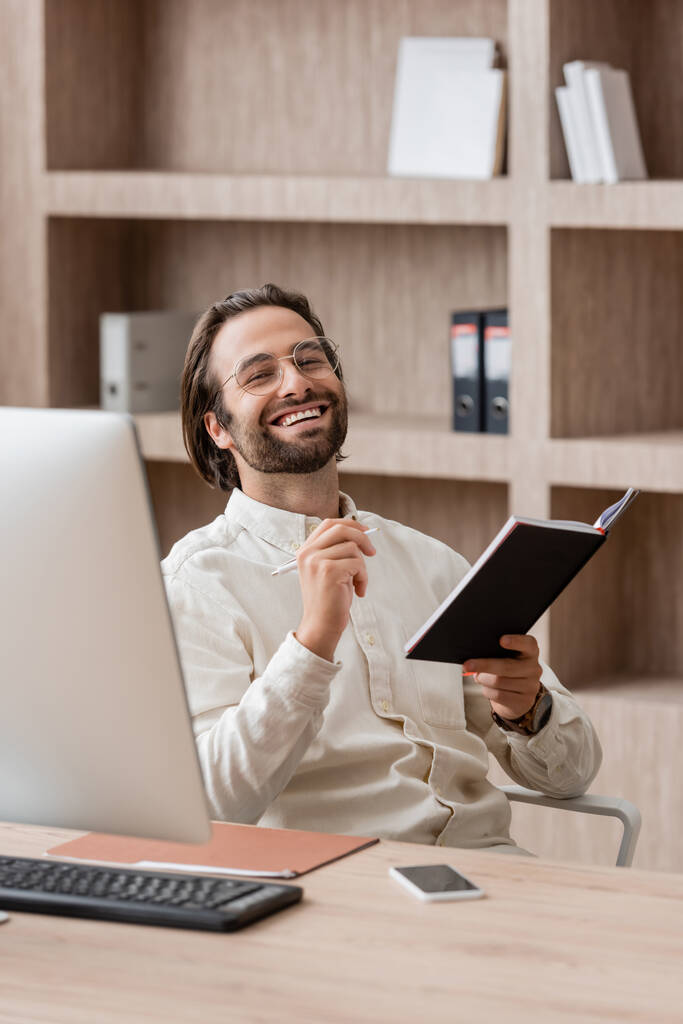 cheerful businessman in eyeglasses holding notepad and smiling at camera near blurred computer monitor - Foto, imagen