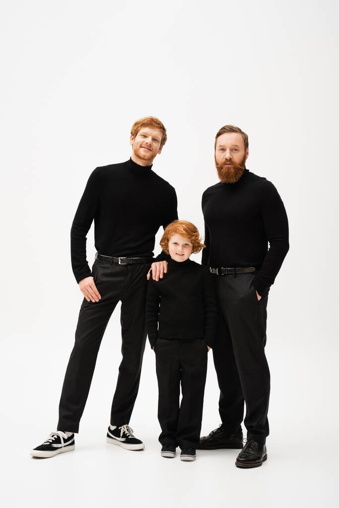 full length of adult bearded men and happy redhead boy standing with hands in pockets and looking at camera on grey background - Photo, image