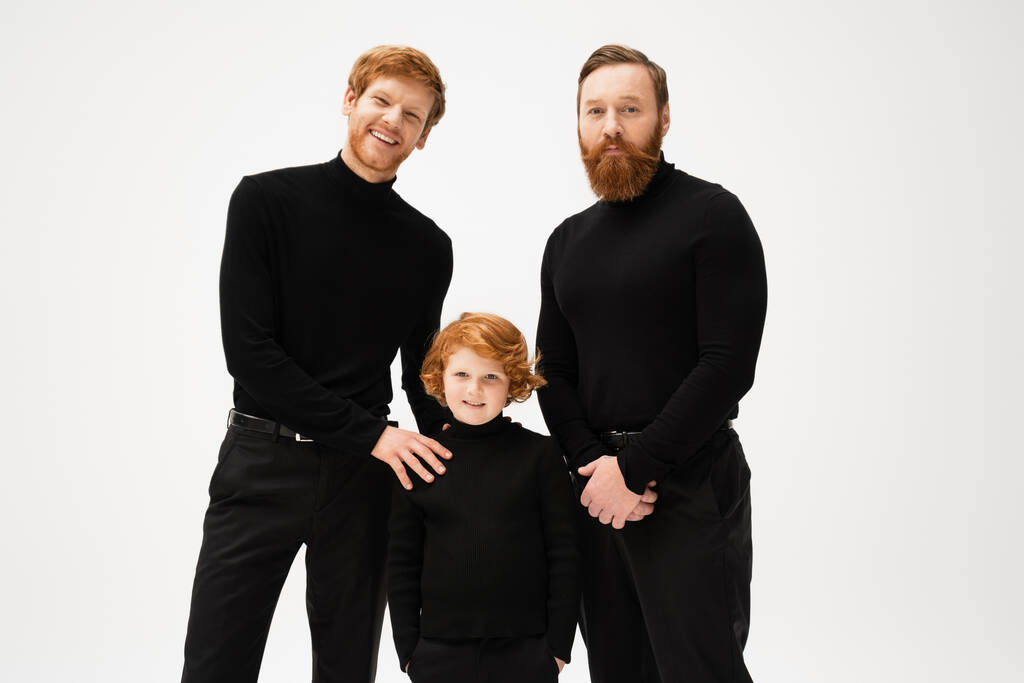 redhead boy standing with hands in pockets near happy dad and grandpa while smiling at camera isolated on grey - Foto, imagen