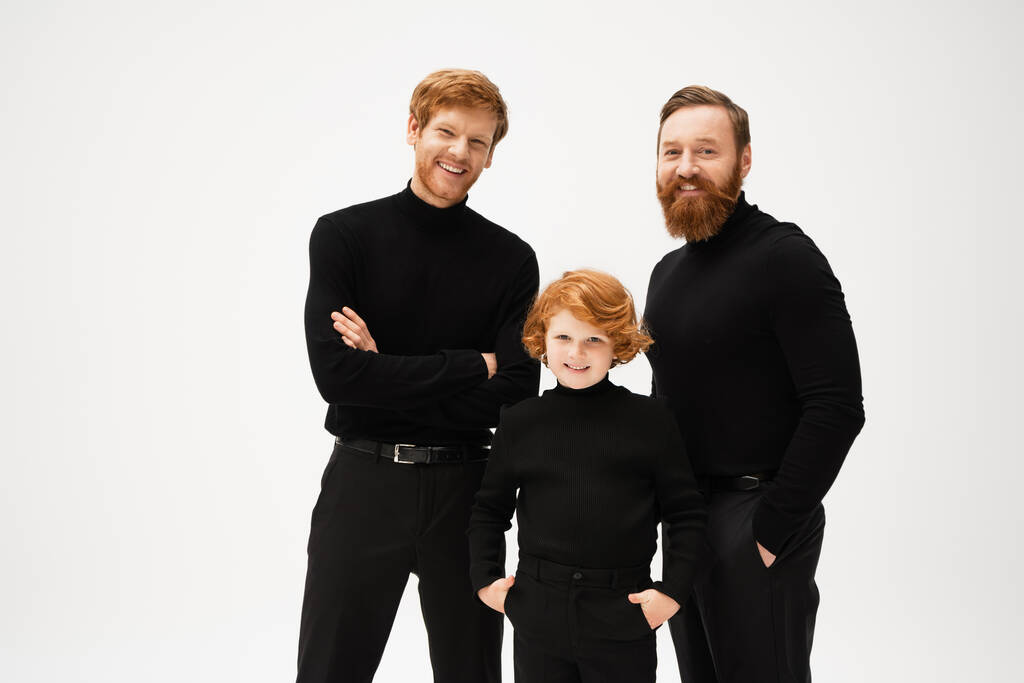 joyful redhead kid standing with hands in pockets near bearded dad and grandfather isolated on grey - Photo, Image