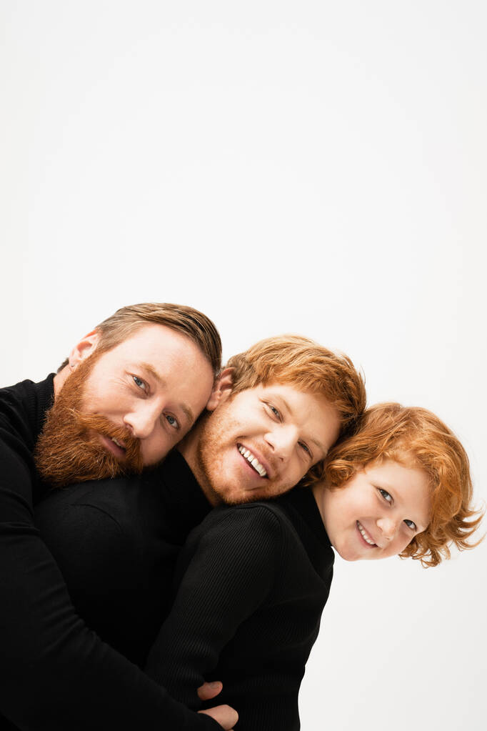 joyful bearded men and redhead kid in black pullovers leaning on each other and smiling at camera isolated on grey - Photo, Image