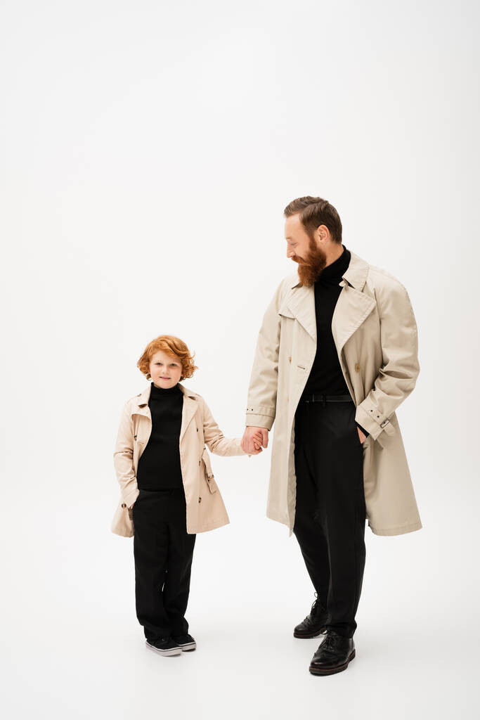 full length of bearded man in trench coat holding hands with redhead grandson posing with hand in pocket on light grey background - Zdjęcie, obraz