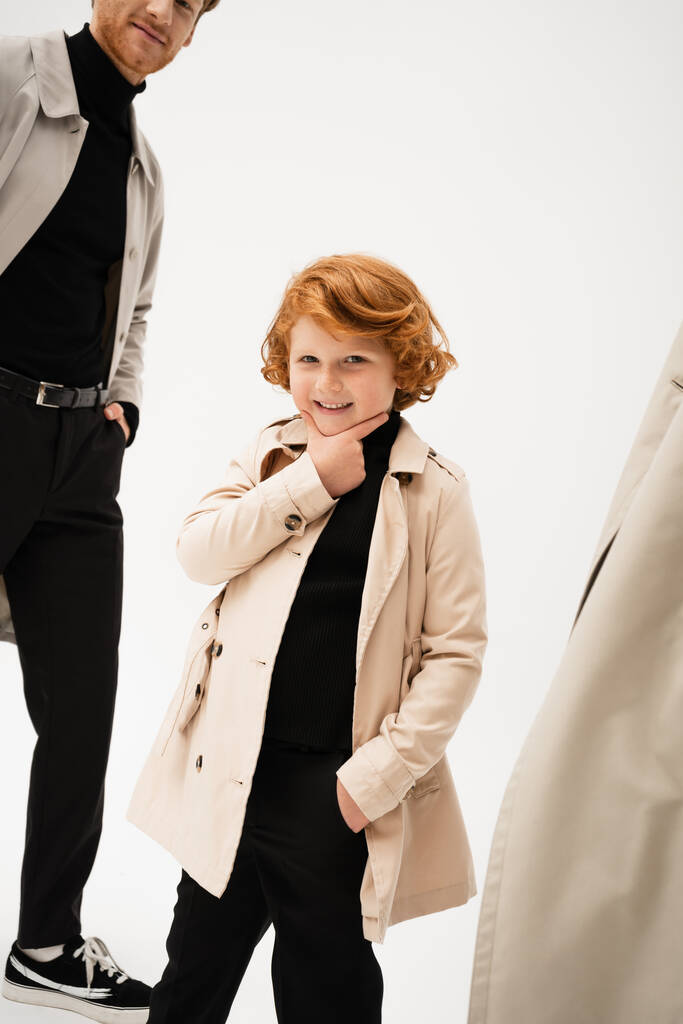 happy redhead boy in trench coat touching chin while posing with hand in pocket on light grey background - Foto, Imagem
