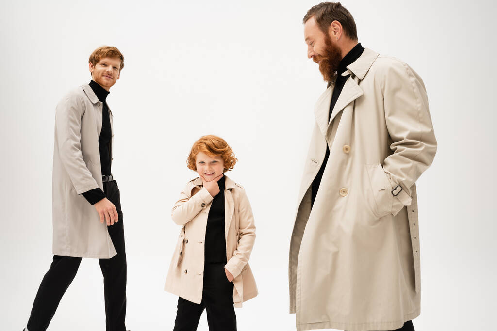 cheerful redhead boy touching chin while posing with hand in pocket near dad and grandpa isolated on grey - Photo, image