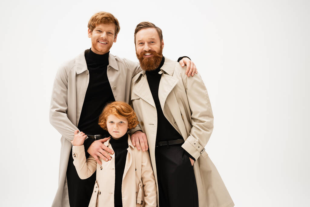 happy bearded men and red haired kid in trench coats looking at camera isolated on grey - Photo, image