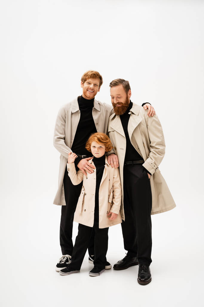 happy redhead man embracing son and bearded father posing in trench coats on light grey background - Foto, Imagem