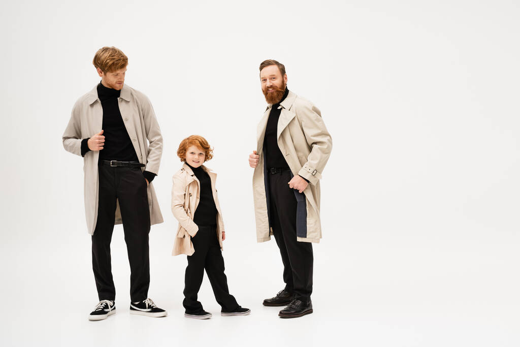 red haired boy smiling at camera while standing with hand in pocket near dad and bearded grandpa in trench coats on light grey background - Foto, Imagem