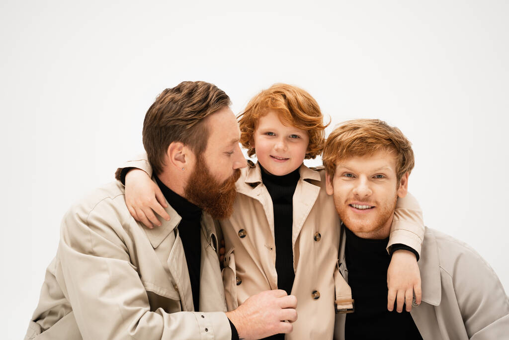 smiling kid in trench coat hugging redhead father and bearded granddad isolated on grey - Foto, Imagen