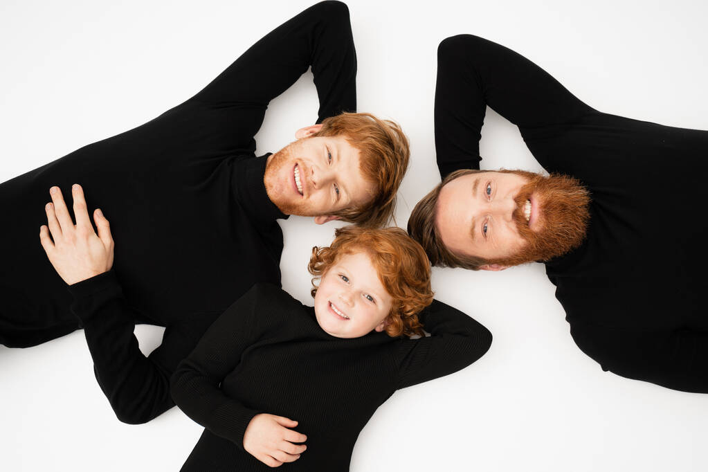 top view of happy redhead men and boy in black sweaters looking at camera while lying on white background - Foto, Bild