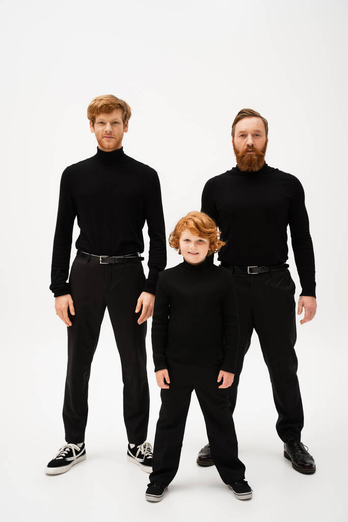 full length of smiling boy and redhead bearded men in black turtlenecks and trousers standing on light grey background - Foto, Imagen