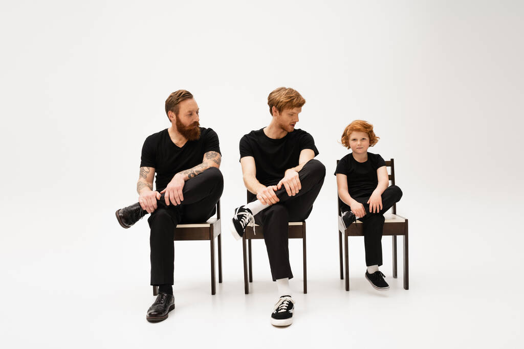 red haired kid looking at camera near father and tattooed grandpa sitting on chairs in black clothes on grey background - Photo, image