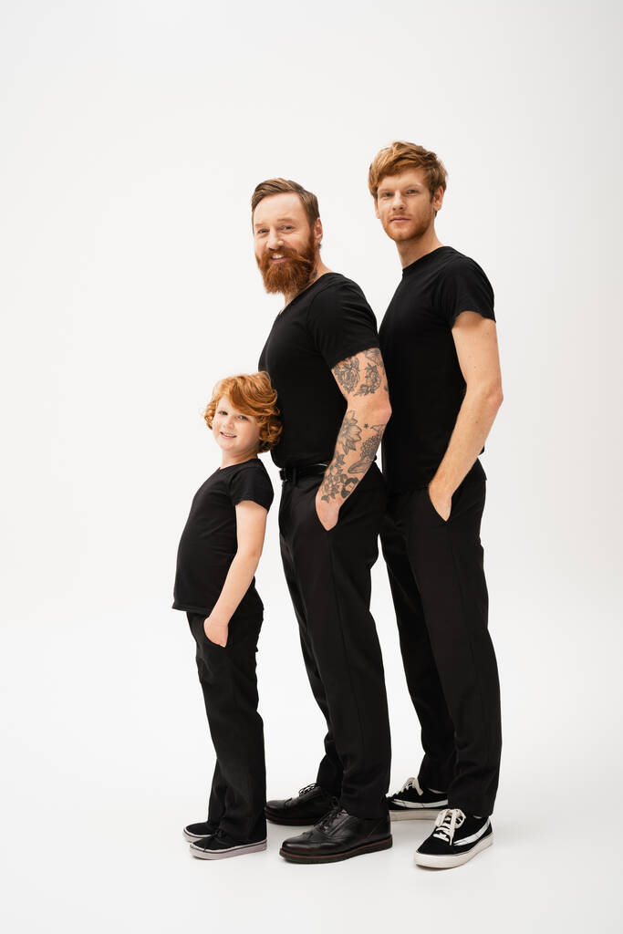 full length of bearded tattooed man with redhead son and grandson posing with hands in pockets of black trousers on light grey background - Foto, Bild