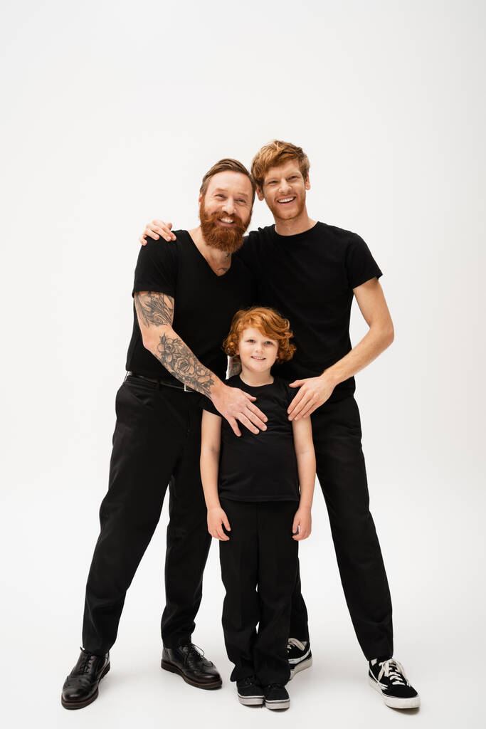 full length of joyful bearded men and redhead kid in black t-shirts and trousers posing on light grey background - Zdjęcie, obraz