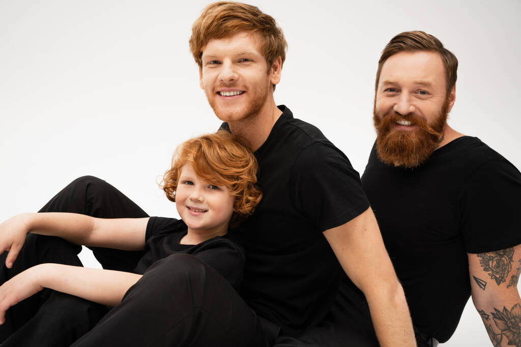 joyful red haired kid with father and bearded grandpa in black t-shirts looking at camera isolated on grey - Fotó, kép