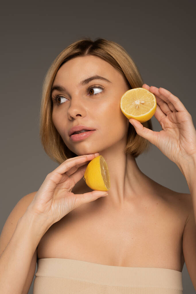 young blonde woman with bare shoulders holding lemon halves and looking away isolated on grey  - Foto, immagini
