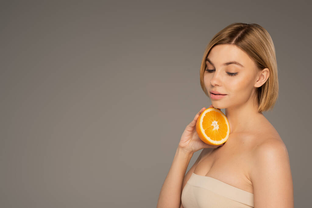pretty young woman with naked shoulders holding orange isolated on grey  - Foto, Imagem