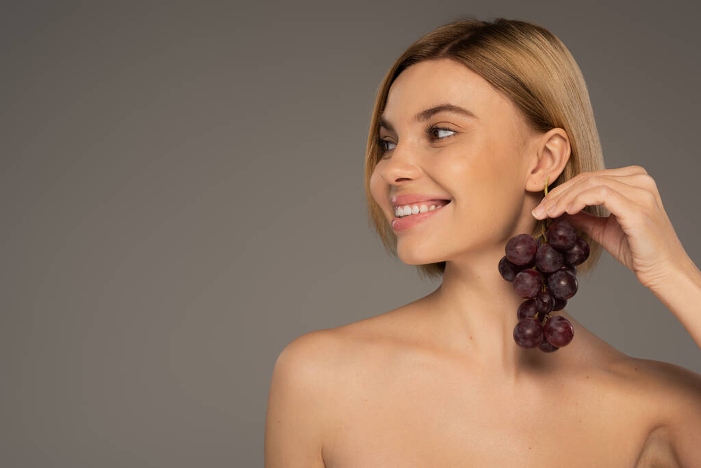 happy young woman with bare shoulders holding ripe grapes near ear isolated on grey  - Photo, Image
