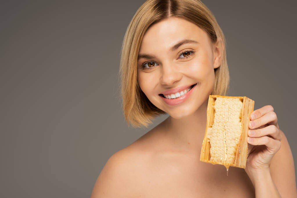 cheerful young woman with bare shoulders holding honeycomb isolated on grey  - Foto, Imagen