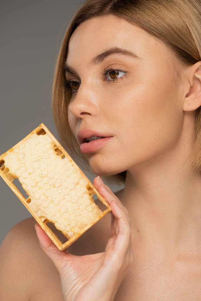 close up of young woman with bare shoulder holding sweet honey in honeycomb isolated on grey - Foto, Bild