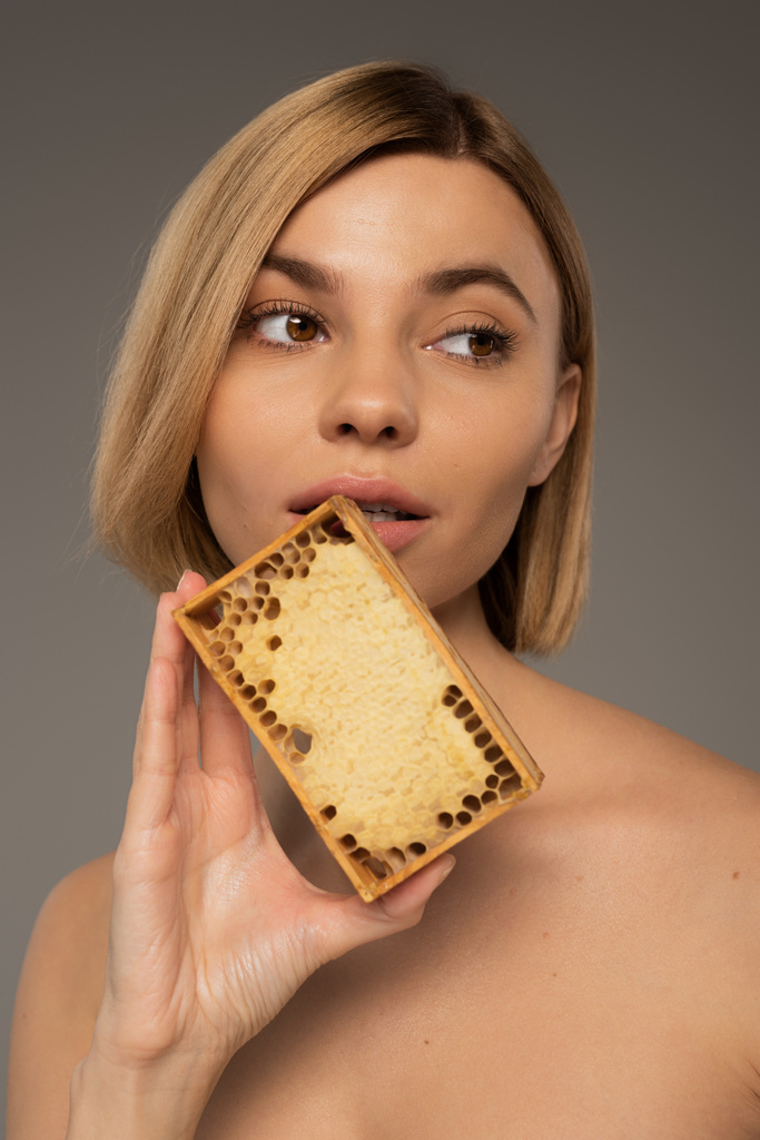 blonde young woman holding wooden frame with sweet honey near face isolated on grey  - Foto, Bild