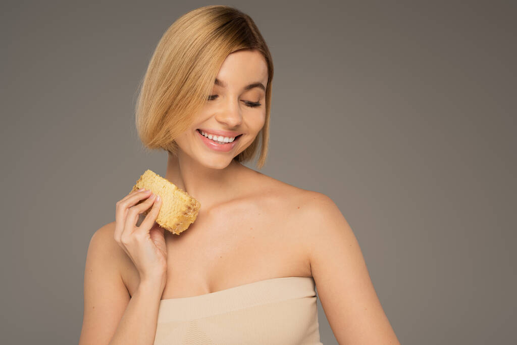 cheerful young woman with bare shoulders holding piece of sweet honeycomb isolated on grey - Foto, immagini
