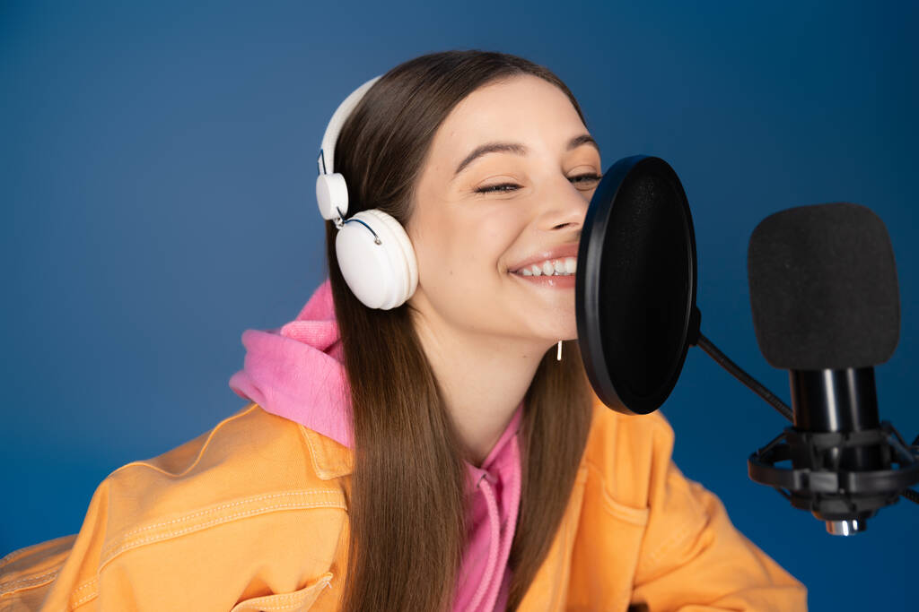 Cheerful teenager in headphones talking near studio microphone during podcast isolated on blue  - Foto, imagen