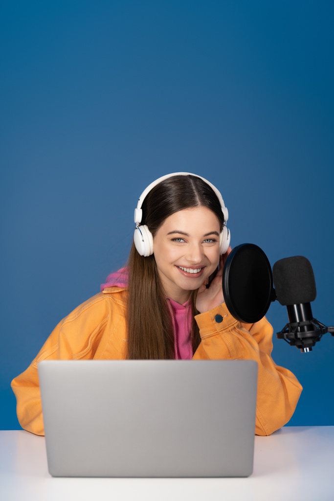 Brunette teen girl in headphones looking at camera near laptop and microphone isolated on blue  - Photo, image