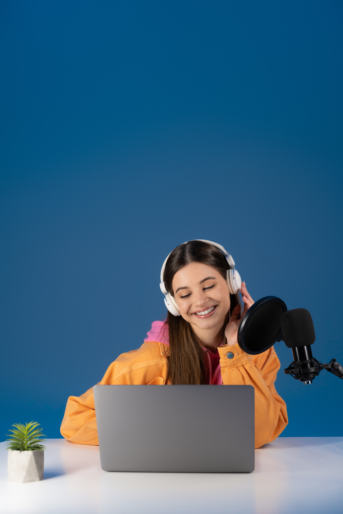 Teen podcaster σε ακουστικά κοιτάζοντας το φορητό υπολογιστή κοντά στο μικρόφωνο στούντιο απομονώνονται σε μπλε  - Φωτογραφία, εικόνα