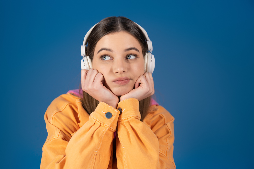 Pensive teenager in wireless headphones looking away isolated on blue  - Foto, afbeelding