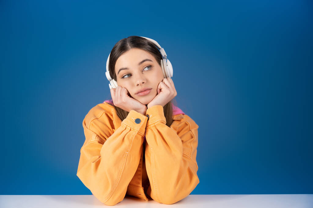 Dreamy brunette teen girl in headphones looking away near table isolated on blue  - Photo, Image