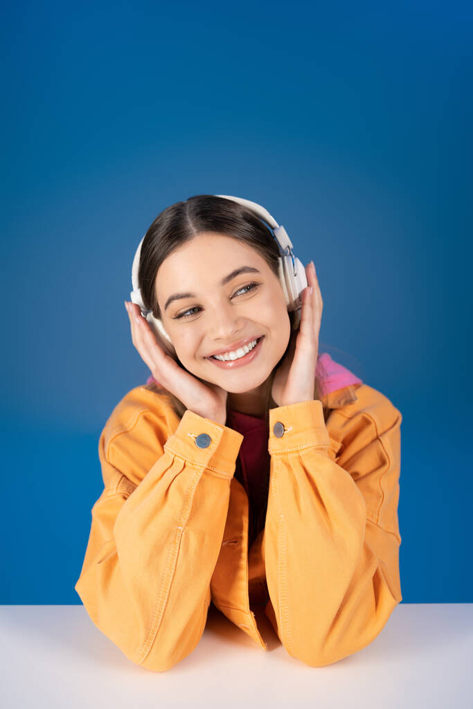 Positive teenager in headphones looking away near table isolated on blue  - Fotó, kép