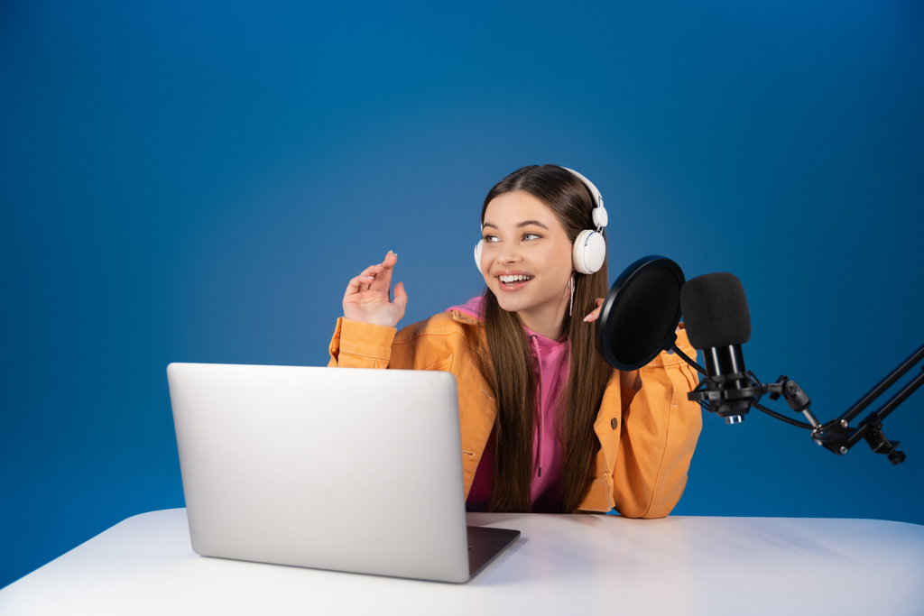 Teen girl in headphones recording podcast near studio microphone and laptop isolated on blue  - Foto, immagini